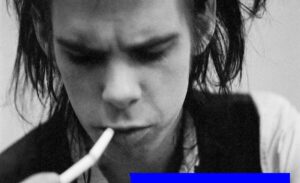 kadr czarno-biały. mężczyzna w ciemnymi półdługimi włosami i papierosem w ustach, pochylony. Nick Cave