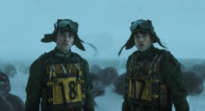 Dwie identyczne postacie (Robert Pattinson) w zielonych skafandrach i goglach stoją na mroźnym, zamglonym terenie. Na ich kombinezonach widnieją duże żółte tabliczki z numerami "18". W tle widać nieokreślone, ciemne kształty.