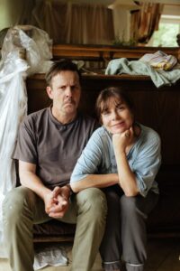 Kadr z filmu „Innego końca nie będzie” ukazuje dwójkę bohaterów siedzących razem na podłodze w zagraconym pomieszczeniu. Mężczyzna z marsową miną ma splecione dłonie, a kobieta uśmiecha się łagodnie, opierając brodę na ręce. Ich ubrania są luźne i robocze, co sugeruje prace domowe lub remont.