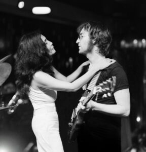 czarno-białe zdjęcie - Yoko Ono i John Lennon. Azjatka w białej sukience trzyma ręce na ramionach mężczyzny w okularach, grającego na gitarze.