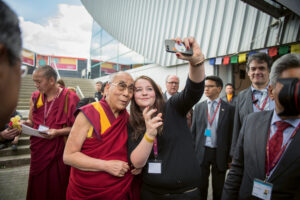 Dalajlama pozuje do selfie z młodą kobietą na tle grupy ludzi i budynku z przeszkloną fasadą. W tle widoczni są mnisi w bordowo-żółtych szatach oraz osoby w garniturach. Na budynku wiszą kolorowe flagi modlitewne.