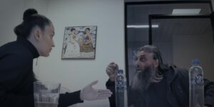 przestrzeń biura. kobieta w koku i czarnym golfie i mężczyzna z długą siwą brodą, tatuażami i długimi włosami rozmawiają, gestykulując