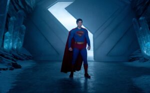kadr. na tle futurystycznej ściany, superman - w niebieskim kombinezomie, czerwonej pelerynie, z literą S na klatce piersiowej.