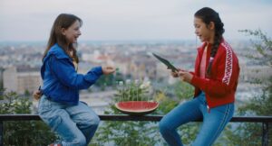 kadr z filmu. Na tle miasta widzianego z oddali, na balustradzie, siedzą dwie nastolatki. Jedna czyta coż z notesu, ma na sobie czerwona kurtkę i dżinsy. Druga ma na sobie niebieską kurtkę i dżinsy, gestykuluje. Miedzy nimi leży arbuz.