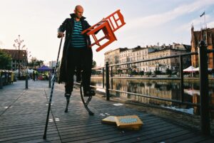 mężczyzna nad rzeką Brdą, trzyma czerwone krzesło, stoiąc na małych szczudłach
