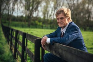 mężczyzna w granatowym garniturze, opiera się o drewniany płot. jest w średnim wieku, ma jasne włosy.