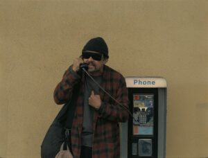 mężczyzna w ciemnych okularach, kraciastej koszuli, czarnej czapce rozmawia przez telefon przy budce telefonicznej wiszącej na żółtej ścianie