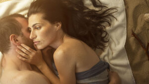 kobieta i mężczyzna, przytulają się. ona zasłania swoją twarzą jego twarz. ma ciemne rozpuszczone włosy. są lekko opiaszczeni.