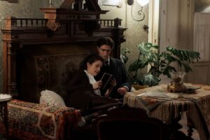 Na zdjęciu widać kobietę i mężczyznę siedzących razem na ozdobnej, drewnianej sofie w elegancko urządzonym, nieco staromodnym pokoju. Kobieta opiera się o mężczyznę i wspólnie czytają książkę. Wokół znajdują się bogato zdobione meble, tkaniny i rośliny doniczkowe, co tworzy przytulną, historyczną atmosferę.