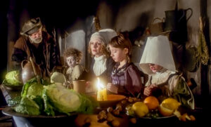 stół bogato zastawiony owocami. przy nim trója dzieci - blady chłopiec z kręconymi włosami, dziewczynka z twarzą częściowo zakrytą białym nakryciem głowy, dziewczynka z jasnymi włosami. Między nimi kobieta w sukni, wpatrzona w przestrzeń, ma kręcone jasne włosy. Nad stołem nachyla się mówiąc coś starszy mężczyzna.