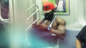 w metrze, muskularny ciemnoskóry mężczyzna, w koszulce, z torbą, w czapce z daszkiem złoto-pomarańczowej, pochyla głowę. nie widać twarzy, jedynie brodę