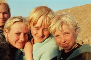 trójka dzieci o blond włosach, opierają się o siebie, z uśmiechem, w tle jasny piach