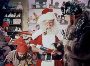 W świątecznym warsztacie Święty Mikołaj trzyma w rękach zabawkę i ma w ustach fajkę. Obok niego stoi osoba w futrzanym stroju, trzymająca mikrofon. W tle pracują elfowie, a półki wypełniają zabawki i lalki