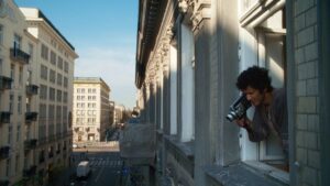 Mężczyzna wychyla się przez okno kamienicy i filmuje ulicę poniżej starą kamerą, na tle szeregu zabytkowych budynków oświetlonych popołudniowym słońcem. kadr z filmu