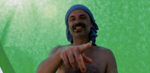 roześmiany mężczyzna z wąsem, bez koszulki, z niebieskim turbanem na głowie, wskazuje palcem w stronę widza, stojąc na zielonym tle.