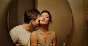 kobieta w kitce, podkoszulku, patrzy przed siebie z lekko przymkniętymi oczyma. mężczyzna w jasnym t-shircie dotyka jej polika i szyi nosem i czołem w geście czułości.