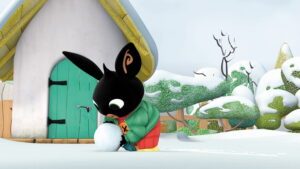 na tle małego domku z zielonymi drzwiami, ośnieżonego krajobrazu, czarny królik lepi bałwana ze śniegu.