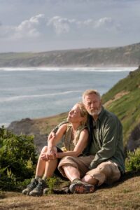 na tle morza i górskiego krajobrazu, przytulona para - dojrzały mężczyzna z brodą, w zielonej koszuli, kobieta z jasnymi włosami, w bluzce bez rękawów.