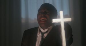 roześmiany ciemnoskóry mężczyzna w marynarce, białej koszuli, z krawatem, przed nim jaśniejący krzyż.