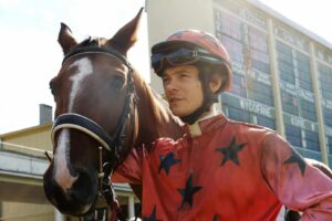 mężczyzna z czerwonym kasku jeździeckim w gwiazdy i bluzie w gwiazdy, trzyma konia, który opiera łeb o jego ramię. w tle budynek, blok.