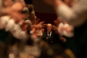 kadr z filmu. starszy, łysiejący mężczyzna w garniturze, otoczony nieostrymi w kadrze klaszczącymi dłońmi.