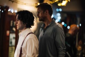 dwóch mężczyzn z koszulach, jeden ma kręcone włosy, drugi nieco starszy i wyższy, ma lekki zarost, stoi za tym młodszym, blisko.