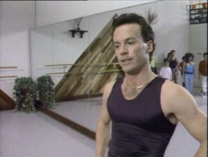 Kadr z sali tanecznej: mężczyzna w czarnym podkoszulku stoi na pierwszym planie, za nim lustra i grupa ćwiczących tancerzy.