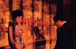 Kobieta w kolorowej, dopasowanej sukni stoi ze spuszczonym wzrokiem przy starej, ceglanej ścianie oklejonej plakatami z chińskimi znakami. Naprzeciw niej, w cieniu, stoi mężczyzna w ciemnym garniturze, pochylony i palący papierosa. Scena utrzymana jest w ciepłych, czerwono-pomarańczowych barwach, z wyraźnymi cieniami, tworząc nastrój intymności i napięcia.