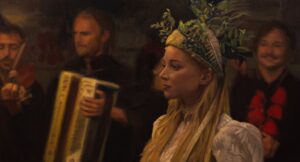 kobieta w sukni, weselnym nakryciu głowy, blondynka, za nią grupa ludzi, w tym akordeonista