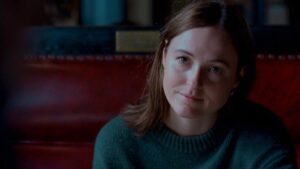 portret szatynki w swetrze w kolorze butelkowej zieleni na tle czerwonego mebla. kobieta ma poważne spojrzenie.