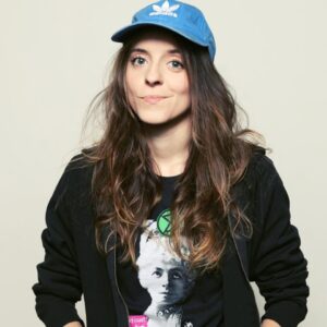 Osoba z długimi brązowymi włosami w niebieskiej czapce z daszkiem i czarnej bluzie, stojąca na jasnym tle. Na koszulce widoczny portret kobiety.