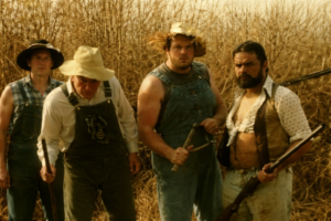 Czterech mężczyzn w roboczych ubraniach stoi na suchym polu, trzymają broń i narzędzia; groźna, napięta atmosfera.