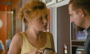 dziewczyna w żółtej bluzce, z rudymi włosami upiętymi w luźny kok, patrzy na mężczyznę z bródką, niewyraźnie widocznego w kadrze w profilu.