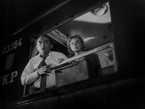 czarno-biały kadr z filmu. w oknie pociągu para - mężczyzna w białej koszuli i kobieta, elegancko ubrana, z półdługimi kręconymi włosami. Patrzą z poważnym wyrazem twarzy przez okno.