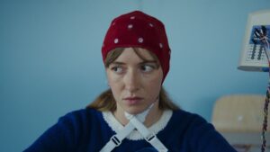 kobieta w czerwonym czepku w białe kropki, z białymi paskami skrzyżowanymi pod szyją, w ciemnym swetrze, patrzy poważnie w lewą stronę kadru