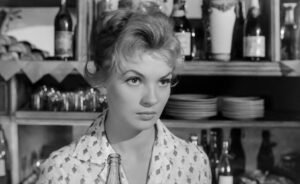 czarno biały kadr. kobieta z upiętymi blond włosami w jasne bluzce we wzorki, na tle półek z naczyniami