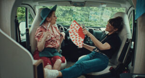 Dwie kobiety siedzą naprzeciw siebie we wnętrzu kampera. Jedna w kapeluszu i wzorzystej bluzce trzyma kieliszek, druga w okularach przeciwsłonecznych wachluje ją papierowym wachlarzem w serduszka. Scena ma luźny, wakacyjny klimat.