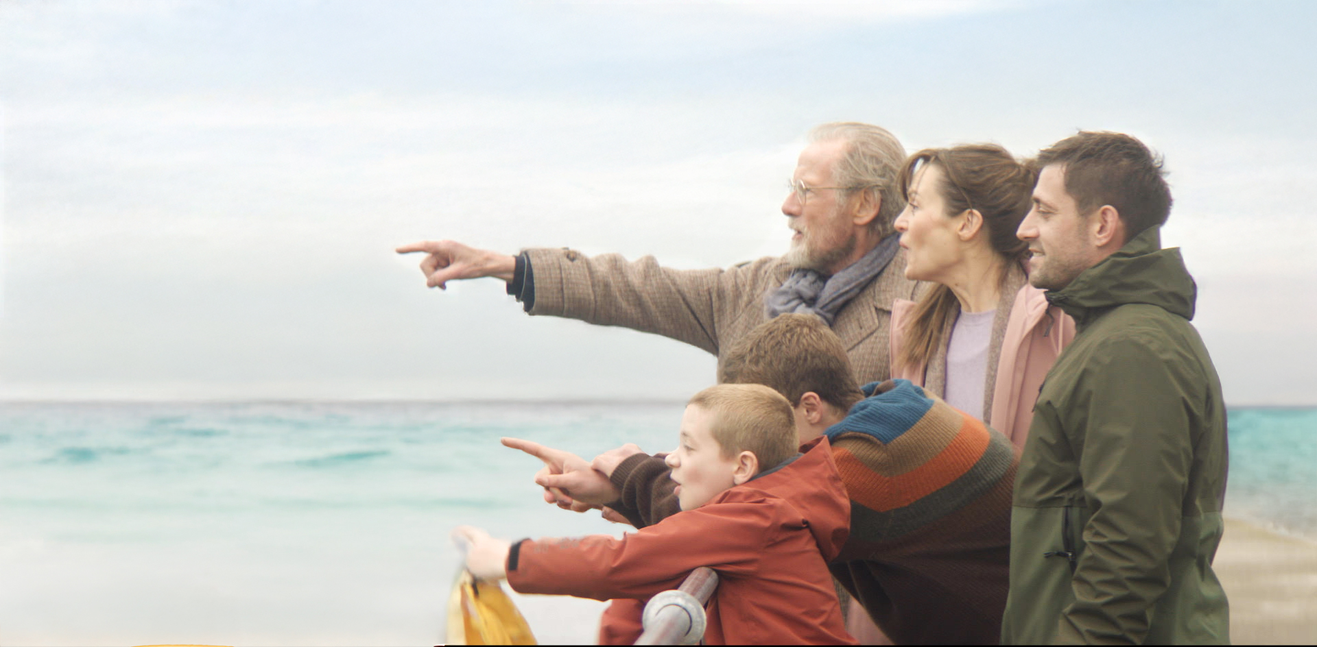 dwóch chłopców, młody mężczyzna i kobieta, starszy mężczyzna, stoją przy barierce i z entuzjazmem wskazują na coś palcami. W tle widać morze.