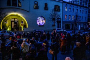 W półmroku tłum zgromadzony przed budynkiem, w oknie wyświetlane są animowane obrazy