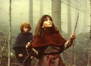 Dwójka dzieci stoi w zamglonym lesie. Na pierwszym planie dziewczynka w brązowej tunice i czerwonej pelerynie trzyma w rękach gałązki i patrzy w górę między drzewa. Za nią stoi chłopiec z kręconymi włosami ubrany w ciemny płaszcz. W tle widać pnie wysokich drzew spowite mgłą.