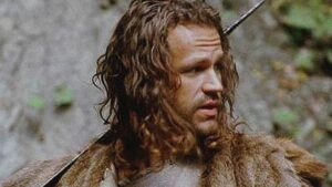 zbliżenie na długowłosego mężczyznę, niosącego na plecach coś w rodzaju szabli. W tle skała