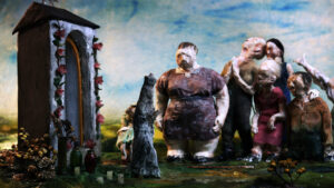 Kadr z filmu animowanego. Grupa zdeformowanych osób stoi przy niewielkiej figurze przedstawiającą Maryję. Przy figurze leżą znicze i zwiędnięte kwiaty,