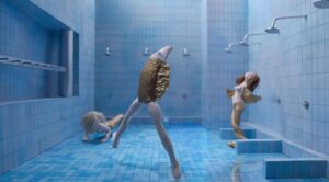 Kadr z filmu animowanego. Wnętrze łazienki w niebieskimi kafelkami na ścianach z dużą ilością pryszniców. Na pierwszym planie ryba z ludzkimi nogami. W tle dwie kobiety z syrenimi ogonami.