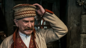 kadr z filmu. mężczyzna z wąsem trzymający dłonią kolorowe wzorzyste nakrycie głowy