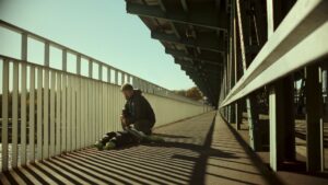 Mężczyzna kuca na długiej, metalowej kładce lub moście, pochylony nad leżącą osobą w śpiworze; ostre światło tworzy rytmiczne cienie barierek na ziemi, a przestrzeń ciągnie się w dal pod konstrukcją.