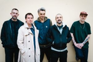 Pięciu dorosłych mężczyzn stoi obok siebie na tle jasnej ściany, ubrani w casualowe kurtki i bluzy, patrzą w stronę aparatu.