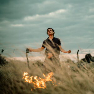 młody mężczyzna z rozłożonymi ramionami, na pierwszym planie płonąca trawa