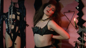 kobieta w czarnej bieliźnie, prowokacyjnie odchylająca głowę, wokół niej łańcuchy, elementy czarnej odzieży, inne dziwnie wyglądające przedmioty