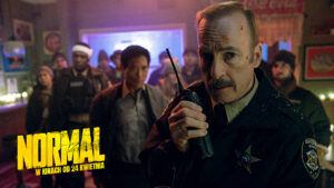 policjant, z wąsem, w mundurze, trzyma krótkofalówkę. za nim grupa ludzi. policjant ma zadrapanie na czole.