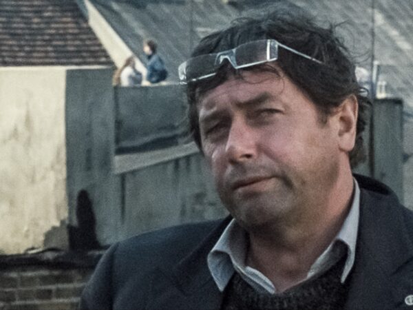 Mężczyzna w średnim wieku stoi na zewnątrz, oparty lub ustawiony blisko metalowej konstrukcji. Ma lekko potargane, ciemne włosy i kilkudniowy zarost. Na głowie ma założone okulary ochronne lub gogle, przesunięte do góry. Ubrany jest w ciemną marynarkę, sweter i koszulę z kołnierzykiem. Jego twarz jest częściowo zmrużona, jakby patrzył w stronę światła. W tle widać przemysłowe otoczenie lub plac budowy, z rozmytą sylwetką osoby na podwyższeniu.